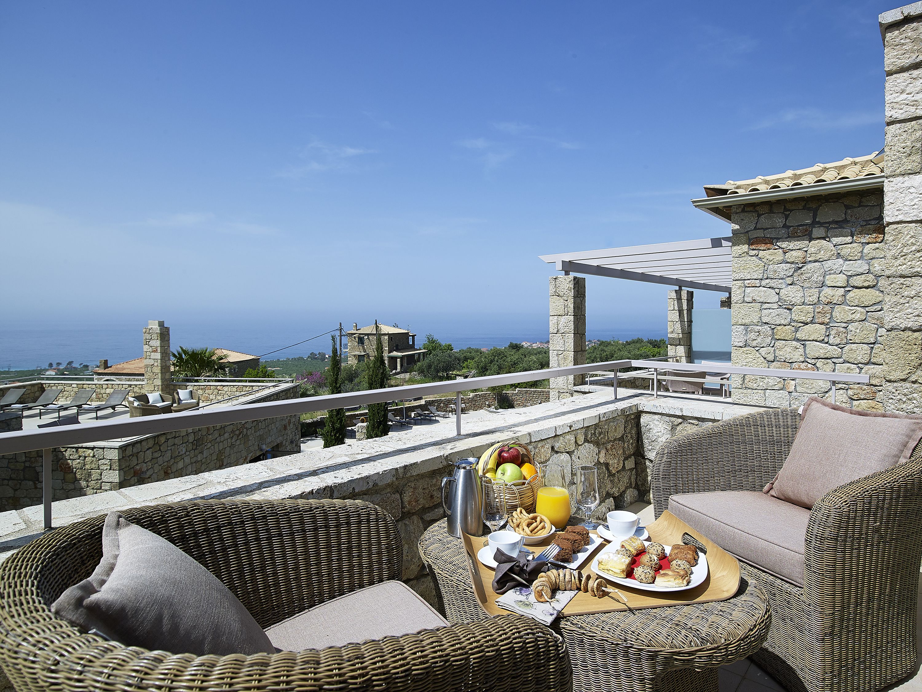 Une terrasse meublée de mobilier en osier, un plateau de petit-déjeuner et un mur de pierre donnant sur une vue imprenable sur la côte.