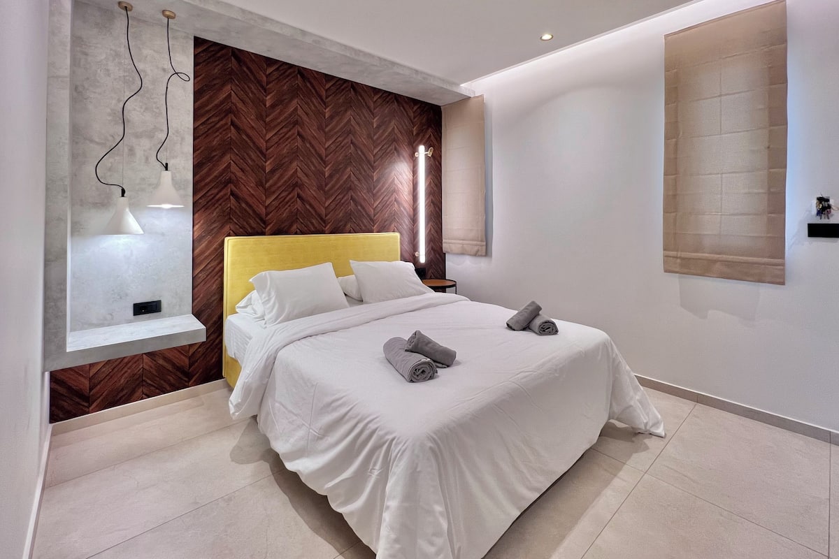 Ein modernes Schlafzimmer mit Bett, weißer Bettwäsche, einer Wand mit Holz-Fischgrätmuster, einer eingebauten Ablage und Pendelleuchten.