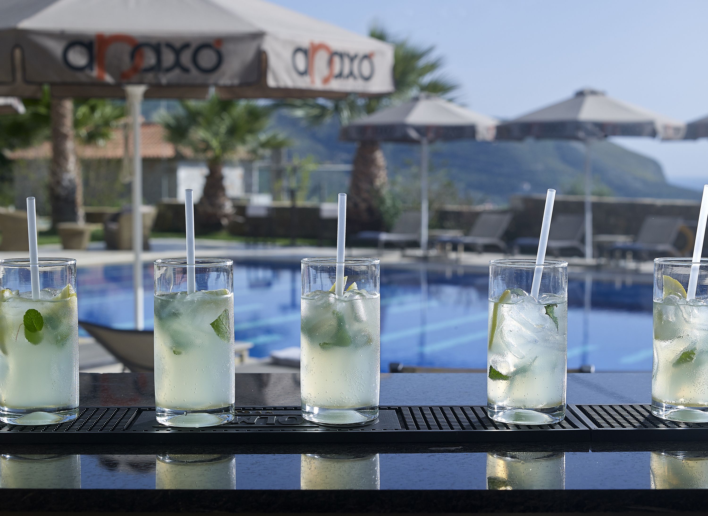 A bar with glasses of a light-colored beverage, mint leaves, and ice, with a pool and umbrellas in the background.
