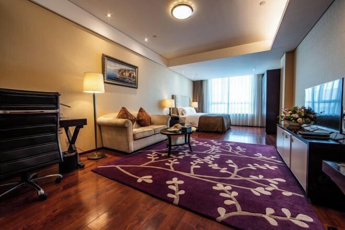 Habitación de hotel con sofá, una mesita, escritorio con computadora, televisor y ventana con cortinas.