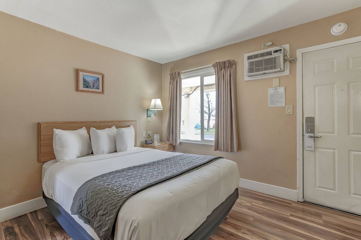 A hotel room with a bed, a window with curtains, an air conditioning unit, a door with a do not disturb sign, and a wall-mounted lamp.