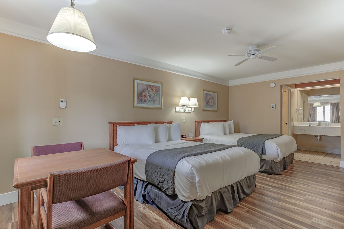 A hotel room with two beds, a wooden table, a chair, a ceiling fan, and a bathroom with a mirror and sink.