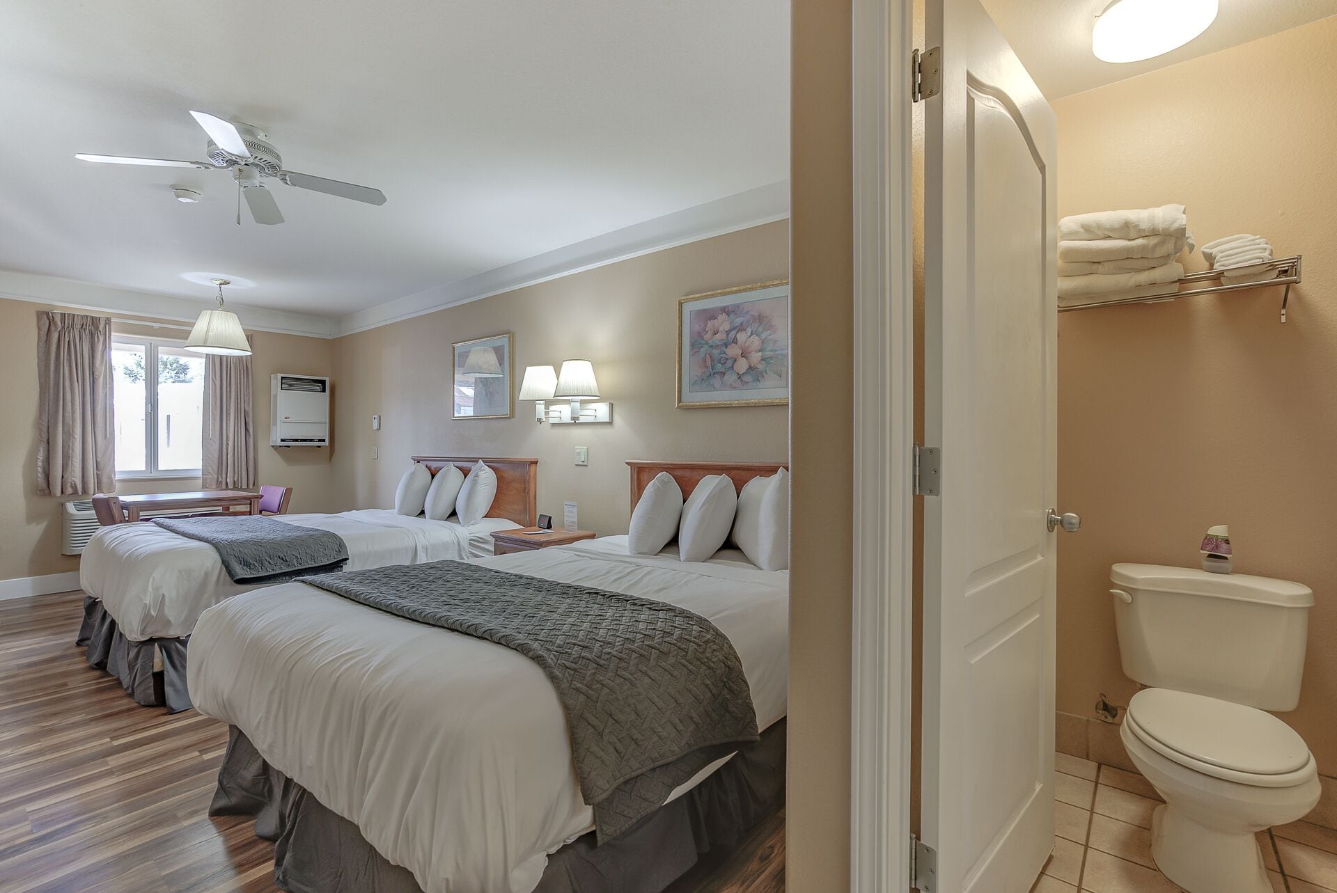 A hotel room with two beds, a ceiling fan, a window with curtains, a bedside table, a lamp, a painting, and a toilet.