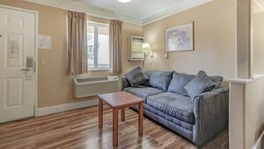 A living room with a grey sofa, a wooden coffee table, a window with curtains, a wall-mounted air conditioner, and a framed picture. - Shasta Pines Motel & Suites (Burney)
