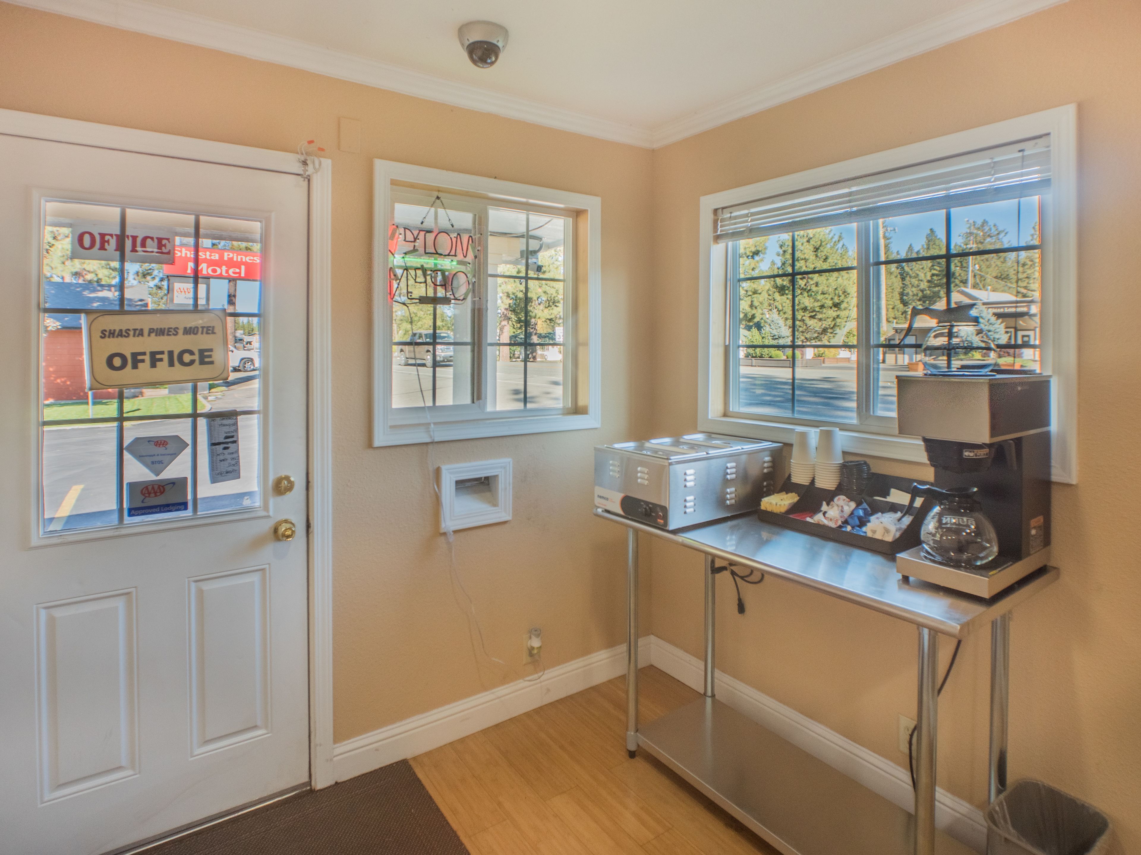 A kitchenette with a coffee maker, a toaster, and a small refrigerator. There is a window with blinds and a door with a sign for the Shasta Pines Motel office.