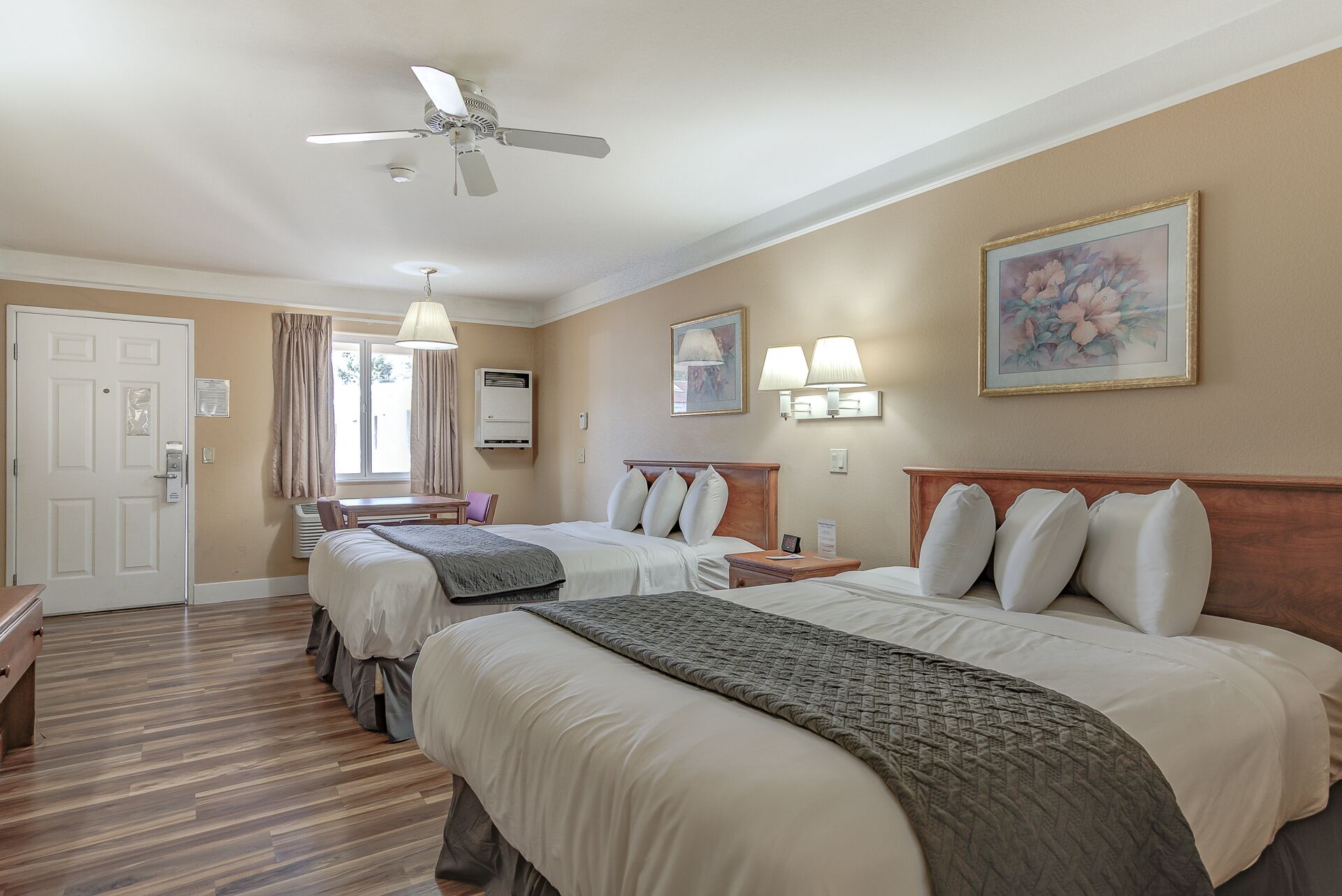 Habitación de hotel con dos camas, ventilador de techo, escritorio, silla, ventana con cortinas y un cuadro en la pared.
