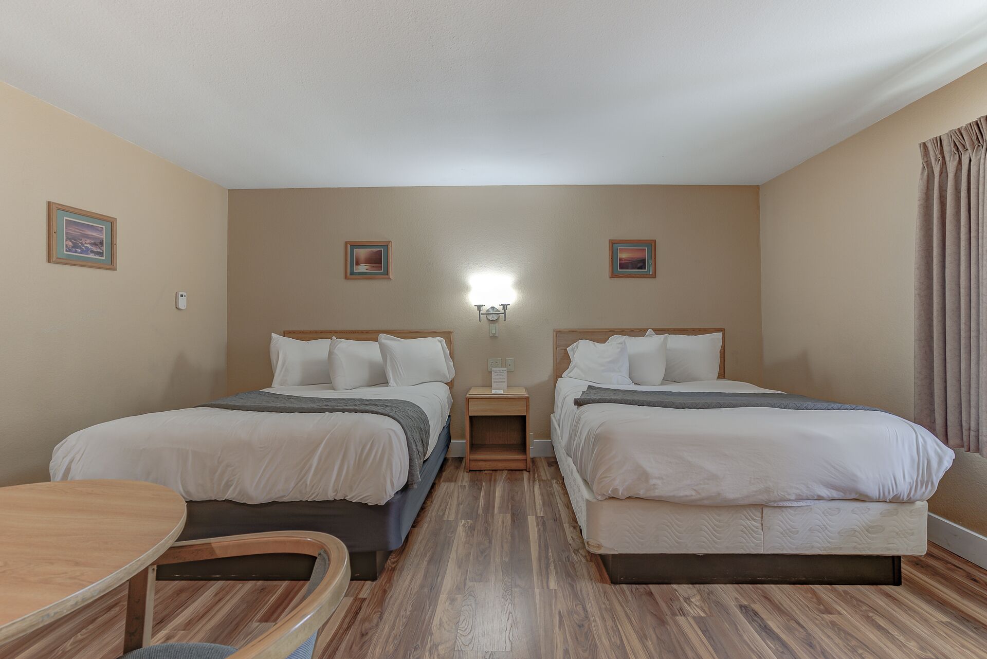 A hotel room with two beds, a small table, and a nightstand with a lamp.