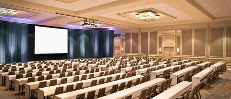 Un salón de conferencias con filas de mesas y sillas, una pantalla grande y piso alfombrado.
