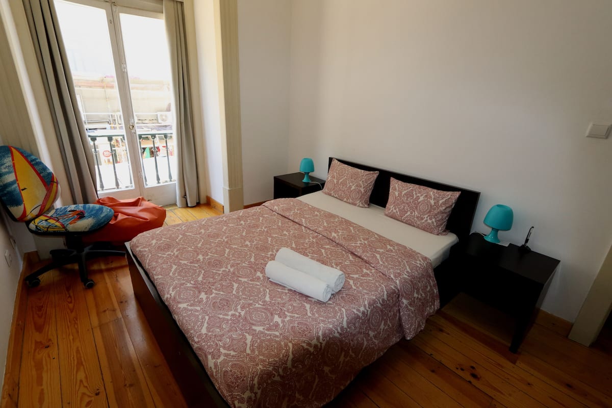Een keurig opgemaakt bed met een beddekleed met dessin, twee nachtlampjes, een stoel met een kleurrijk kussen, en uitzicht buiten door een raam.