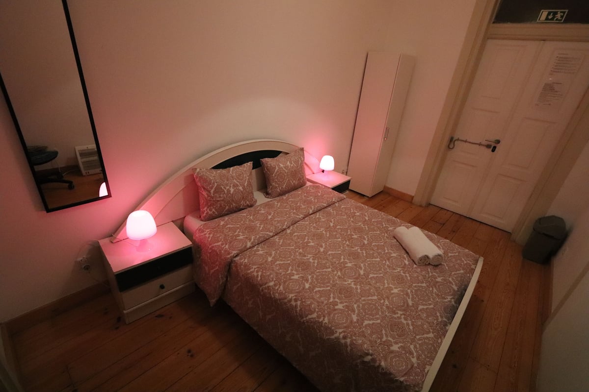 Een slaapkamer met een bed, twee bedlampen, een nachtkastje, een spiegel, een deur en een radiator.