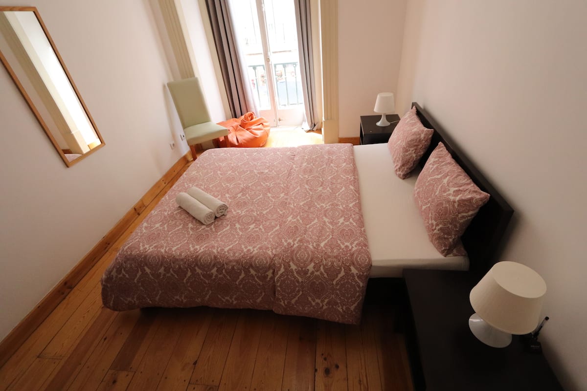 Een slaapkamer met een bed, nachtkastje, lamp, stoel en een raam met gordijnen.