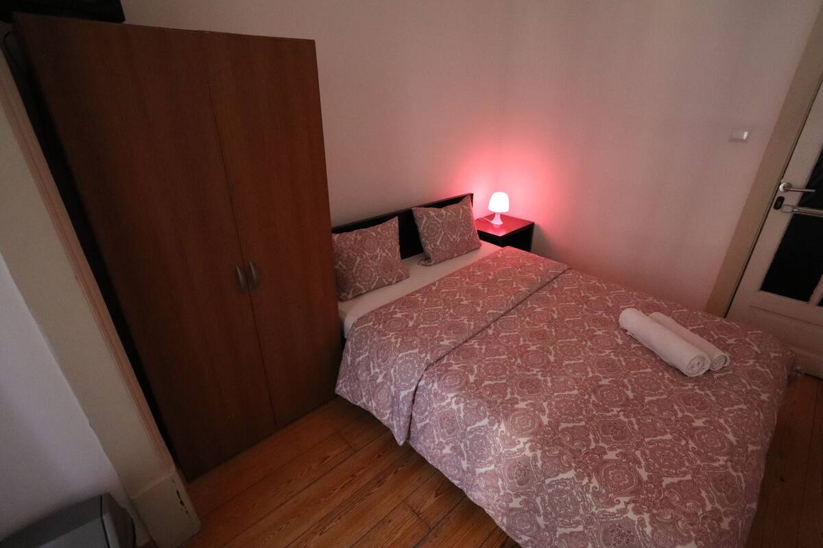 Een slaapkamer met een bed, een nachtkastje met lamp, een houten garderobe en een deur.