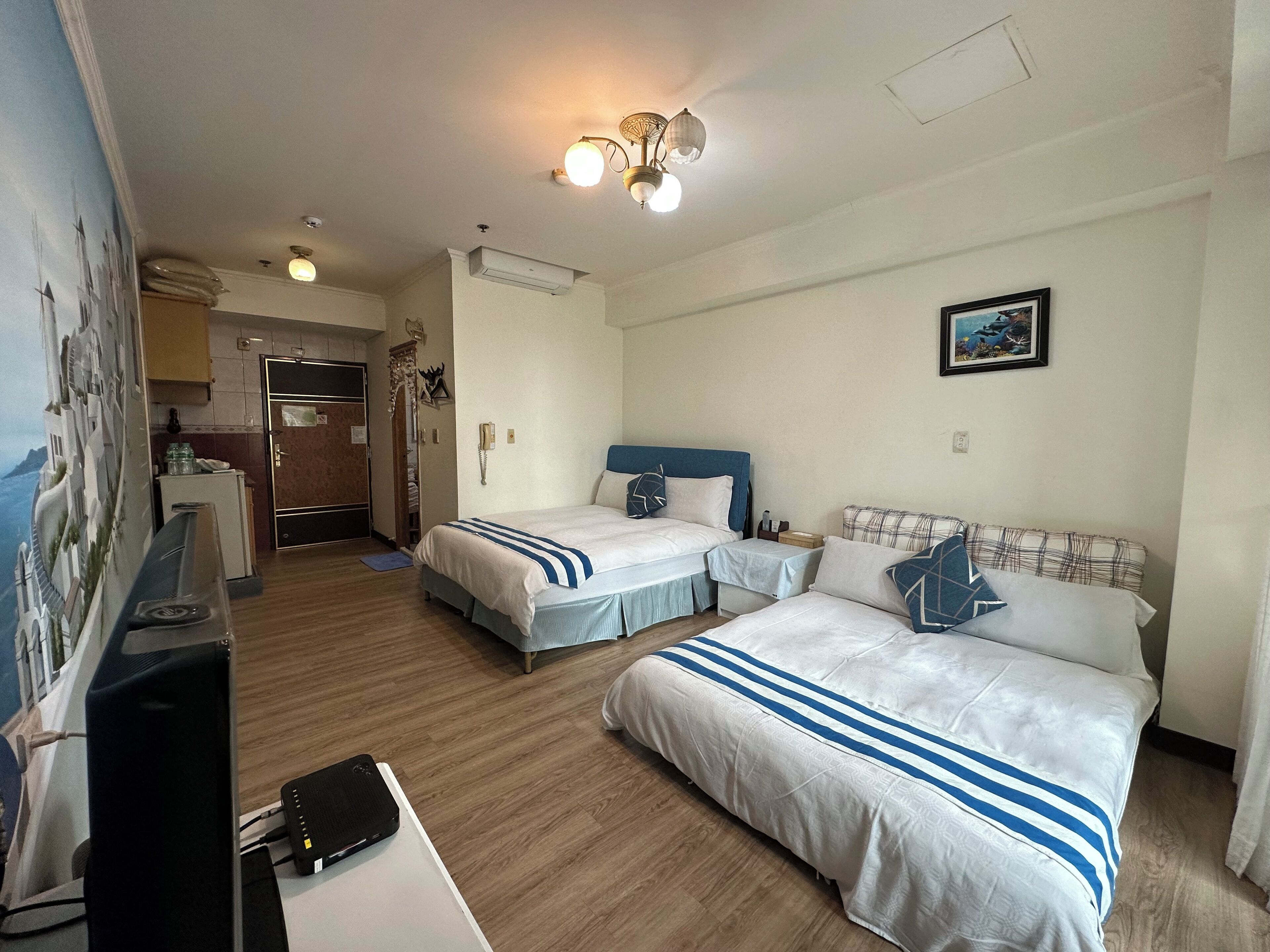 Habitación de hotel con dos camas, un escritorio con computadora, teléfono y una pequeña mesa. Se observa una kitchenette al fondo.
