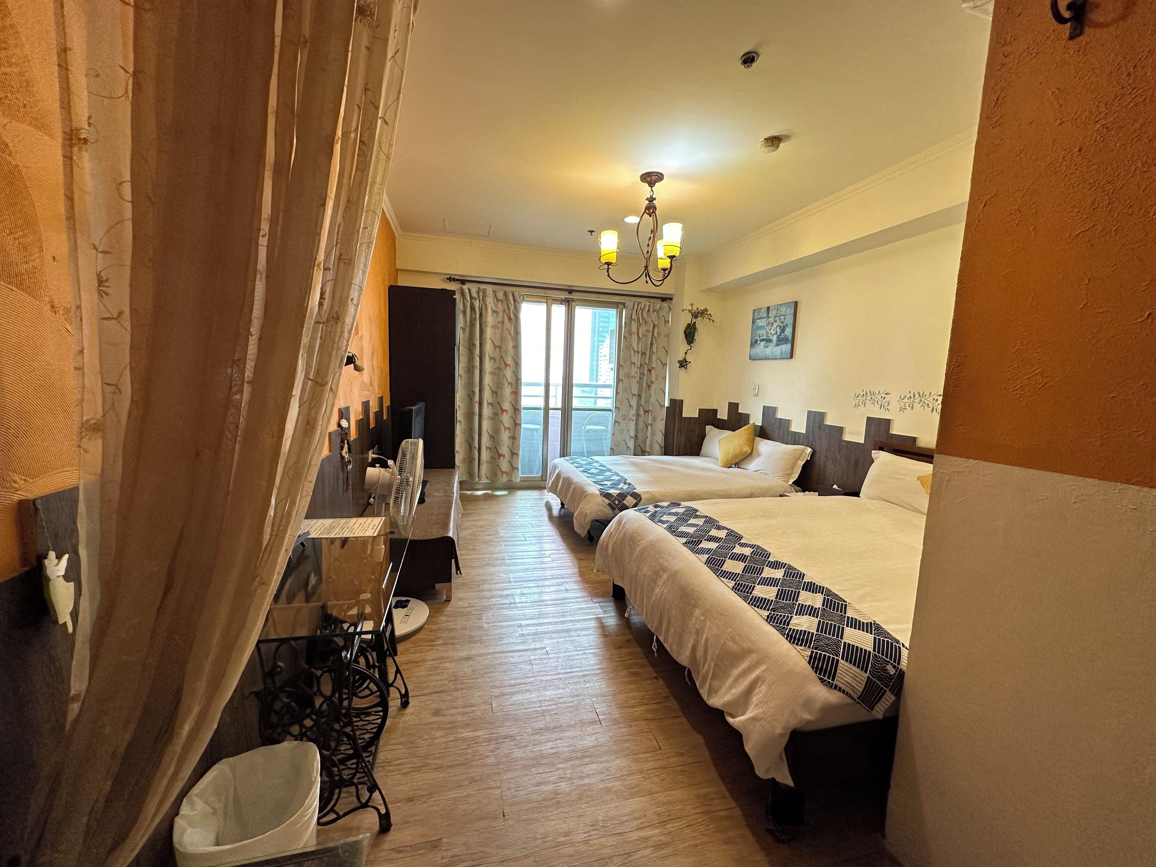 A hotel room with two beds, a desk, a chair, a fan, and a view of the outside.