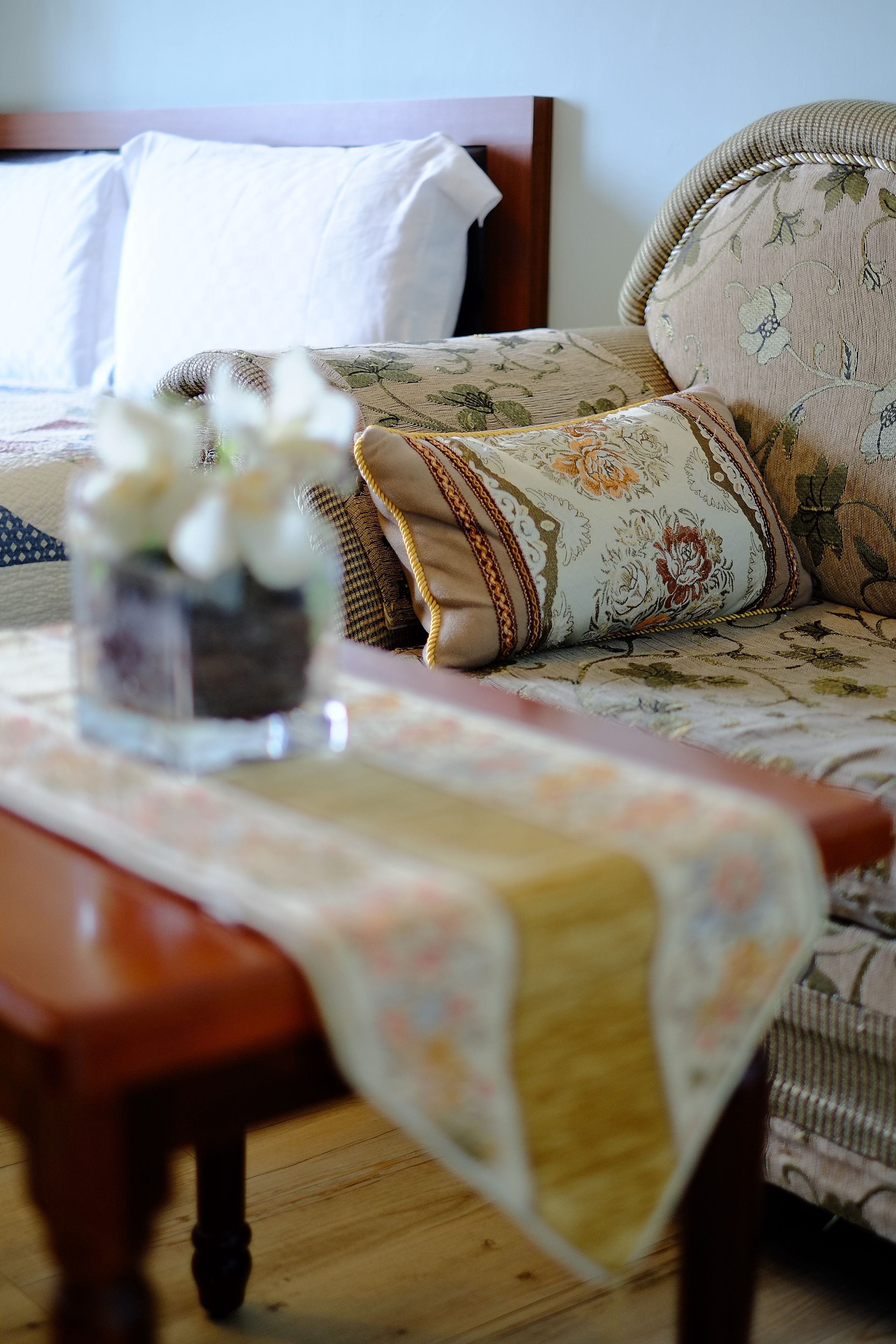 Un dormitorio ordenado con una cama, una mesita de noche con un florero y un sofá estampado con motivos florales.