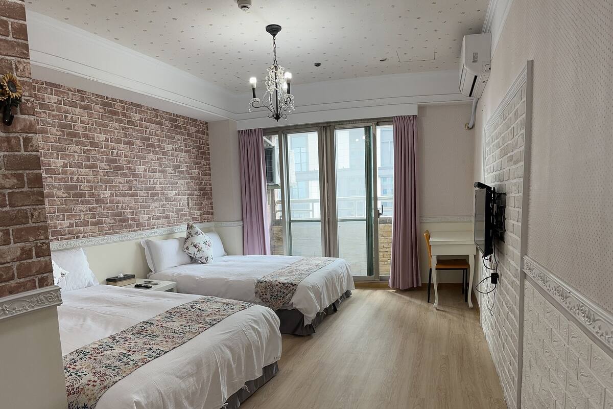 A hotel room with two beds, a chandelier, a TV mounted on the wall, and a balcony with a view.