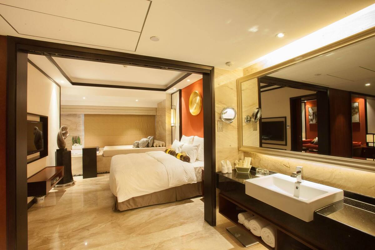 Una habitación de hotel moderna con una cama grande, un sofá independiente, un baño con un espejo grande y un tocador con lavamanos.