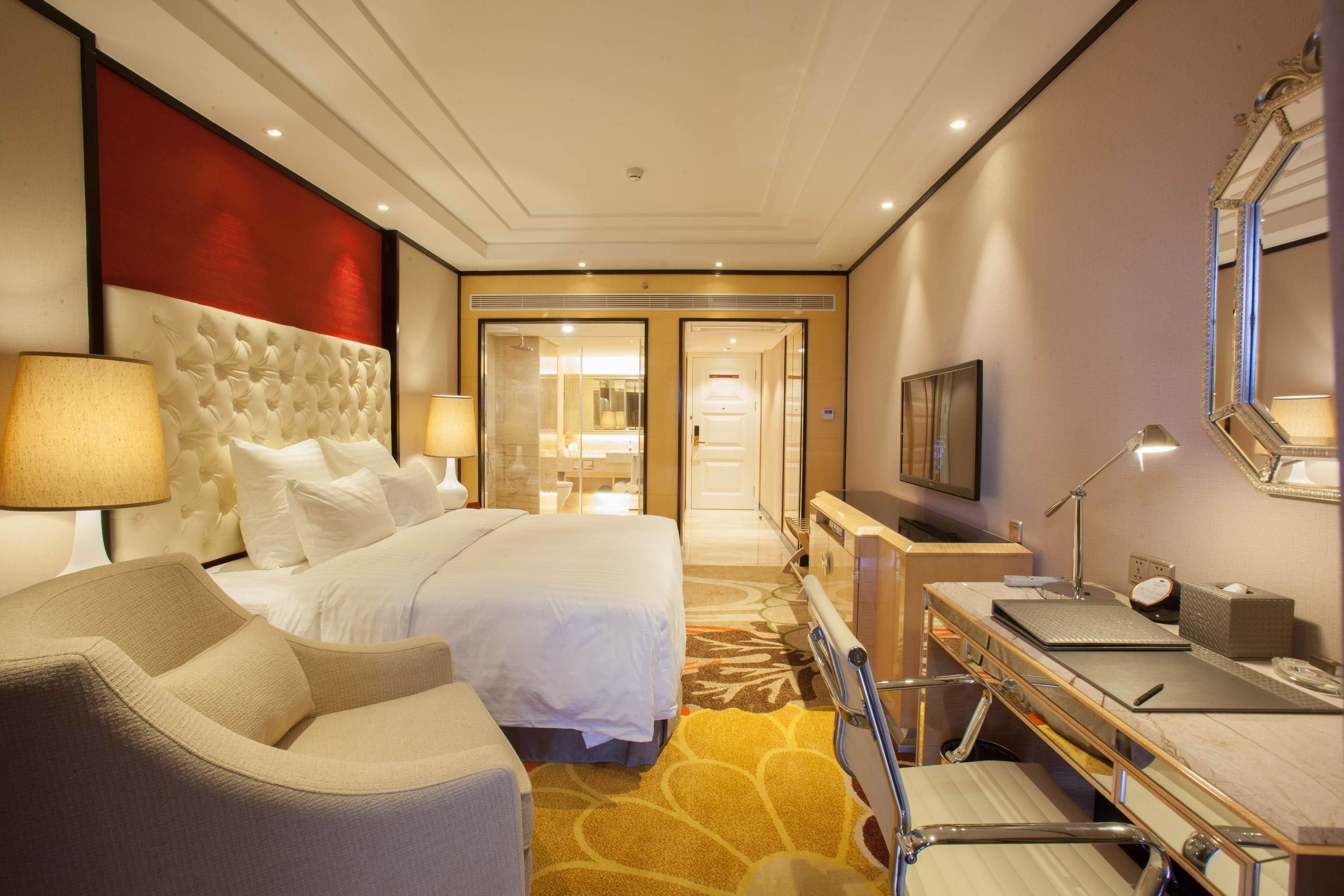 A hotel room with a large bed, a desk with a computer, a chair, a TV, and a bathroom visible through an open door.