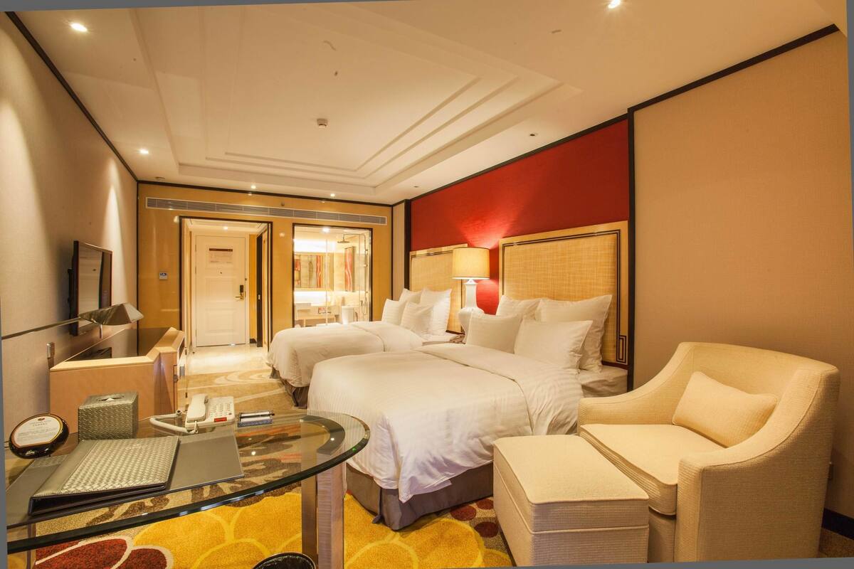 Habitación de hotel con dos camas, un área de descanso con una mesa de vidrio, un escritorio con una computadora y una pared de acento roja.