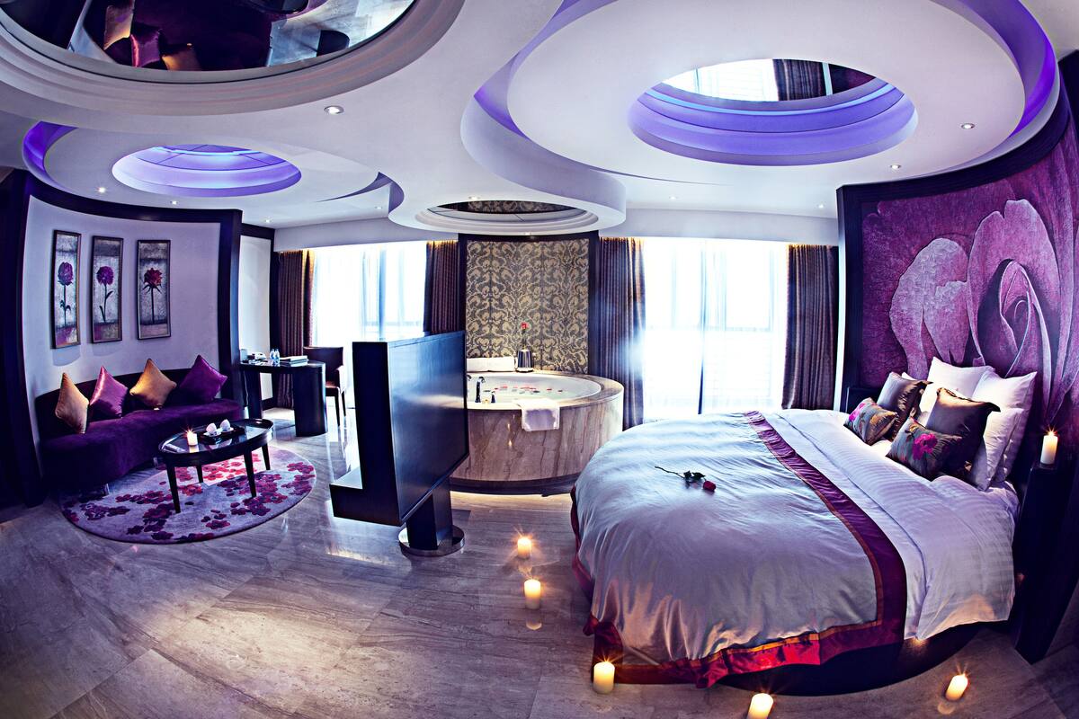 Una habitación de hotel moderna con una cama grande, un área de estar con sofá y mesa de centro, un espacio de baño circular y un amplio ventanal que proporciona luz natural.