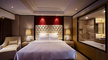 A hotel room with a large bed, a sitting area with a chair, a nightstand with lamps, and a bathroom with a bathtub and mirror.