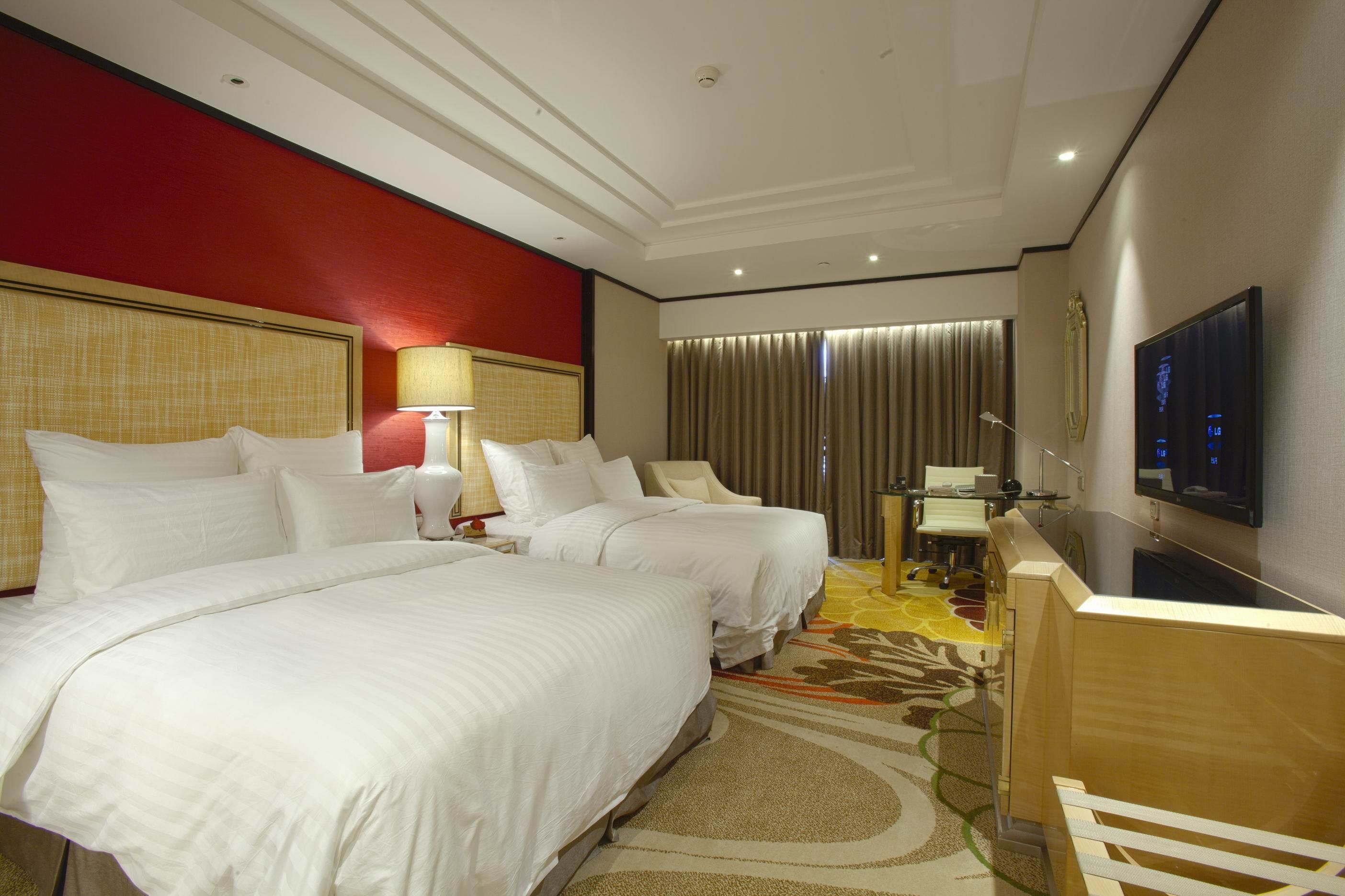 A hotel room with two beds, a desk, a TV, and a red accent wall.