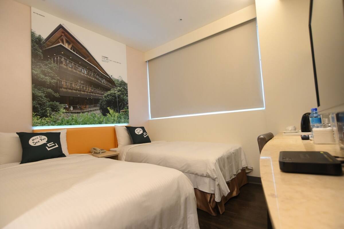 A hotel room with two beds, a desk, a window with blinds, and a poster on the wall.