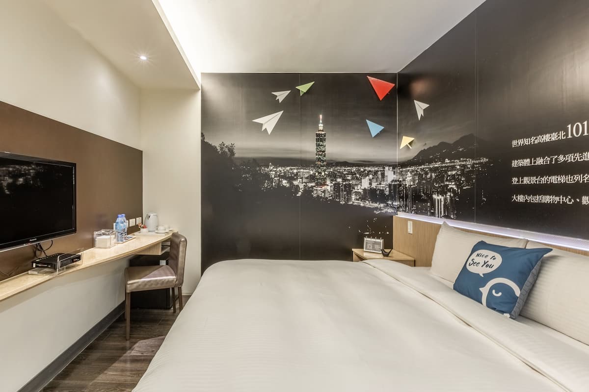 A hotel room with a bed, a desk, a chair, a TV, and a wall with a cityscape and paper airplanes.
