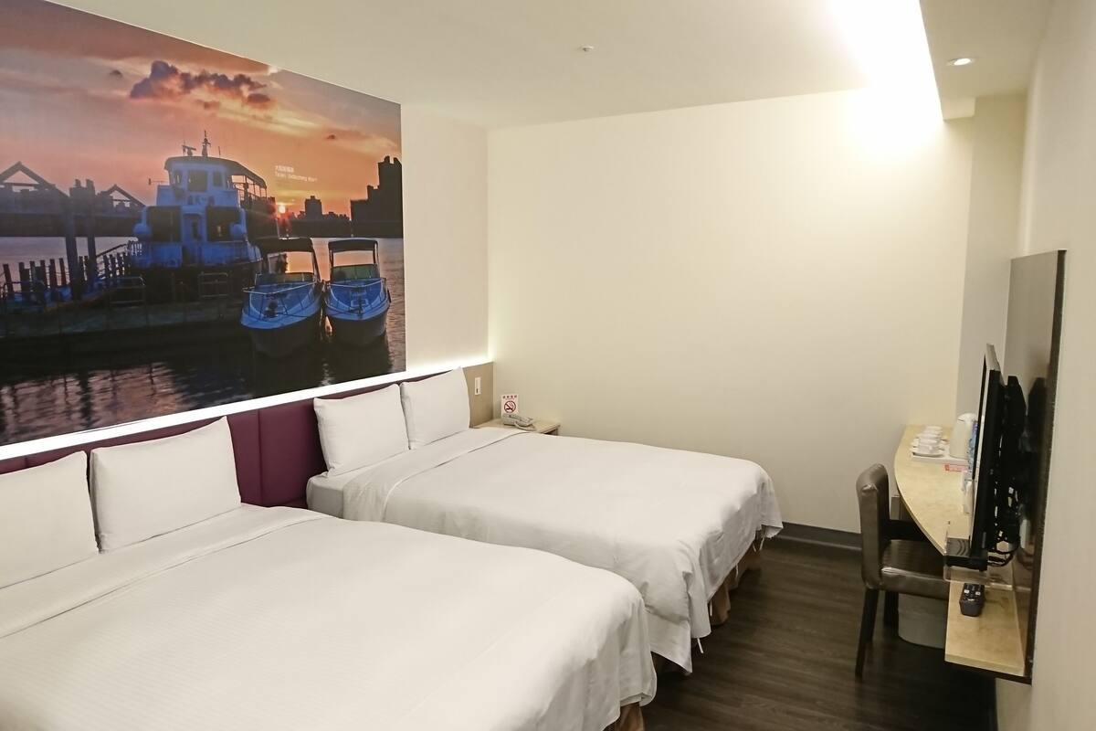 A hotel room with two beds, a painting of boats on water, a desk with a chair, and a TV.