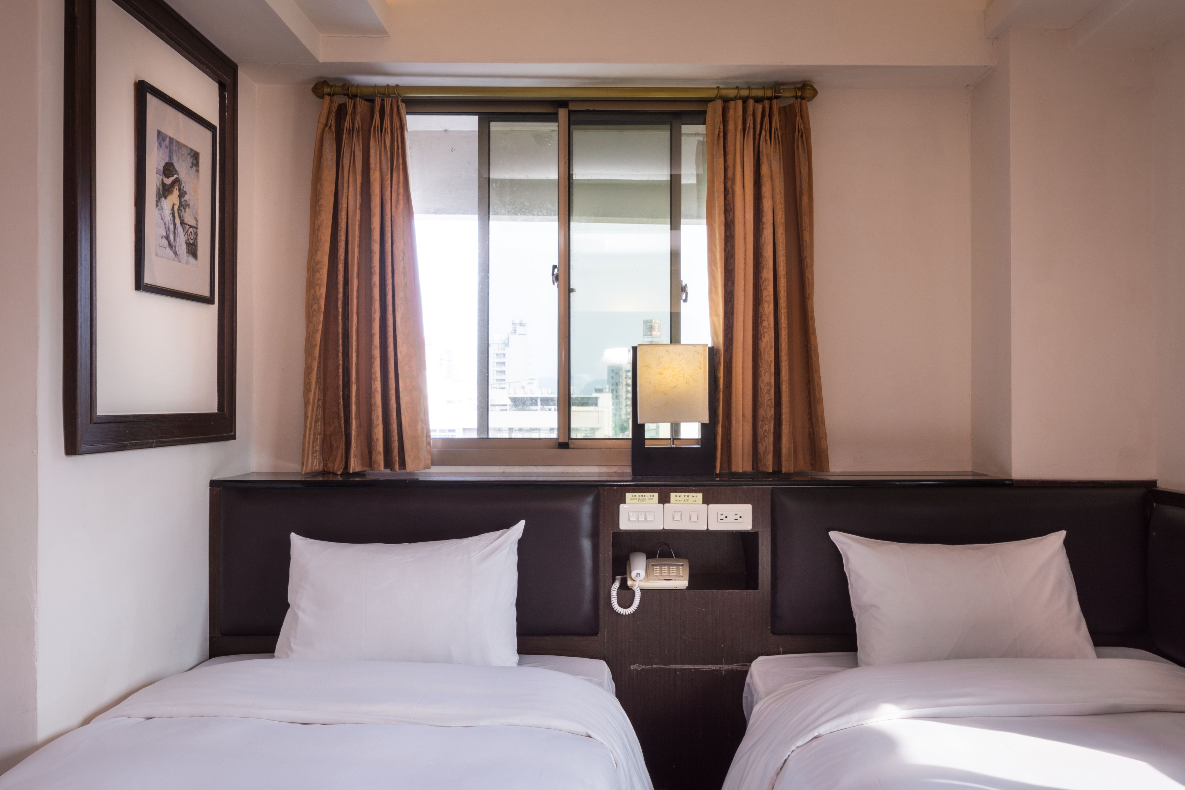A hotel room with two beds, a window with curtains, a framed picture on the wall, and a bedside table with a lamp and phone.