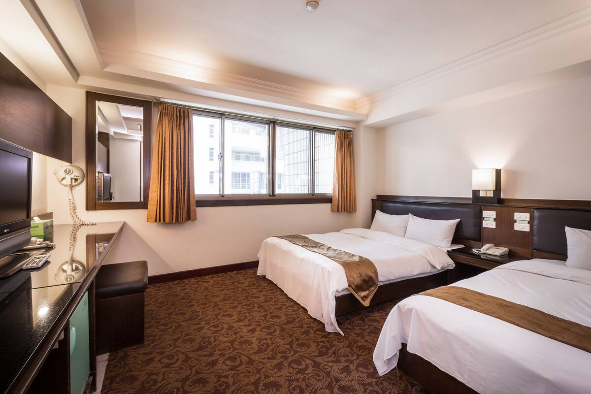 A hotel room with two beds, a desk with a telephone, and a large window with curtains.