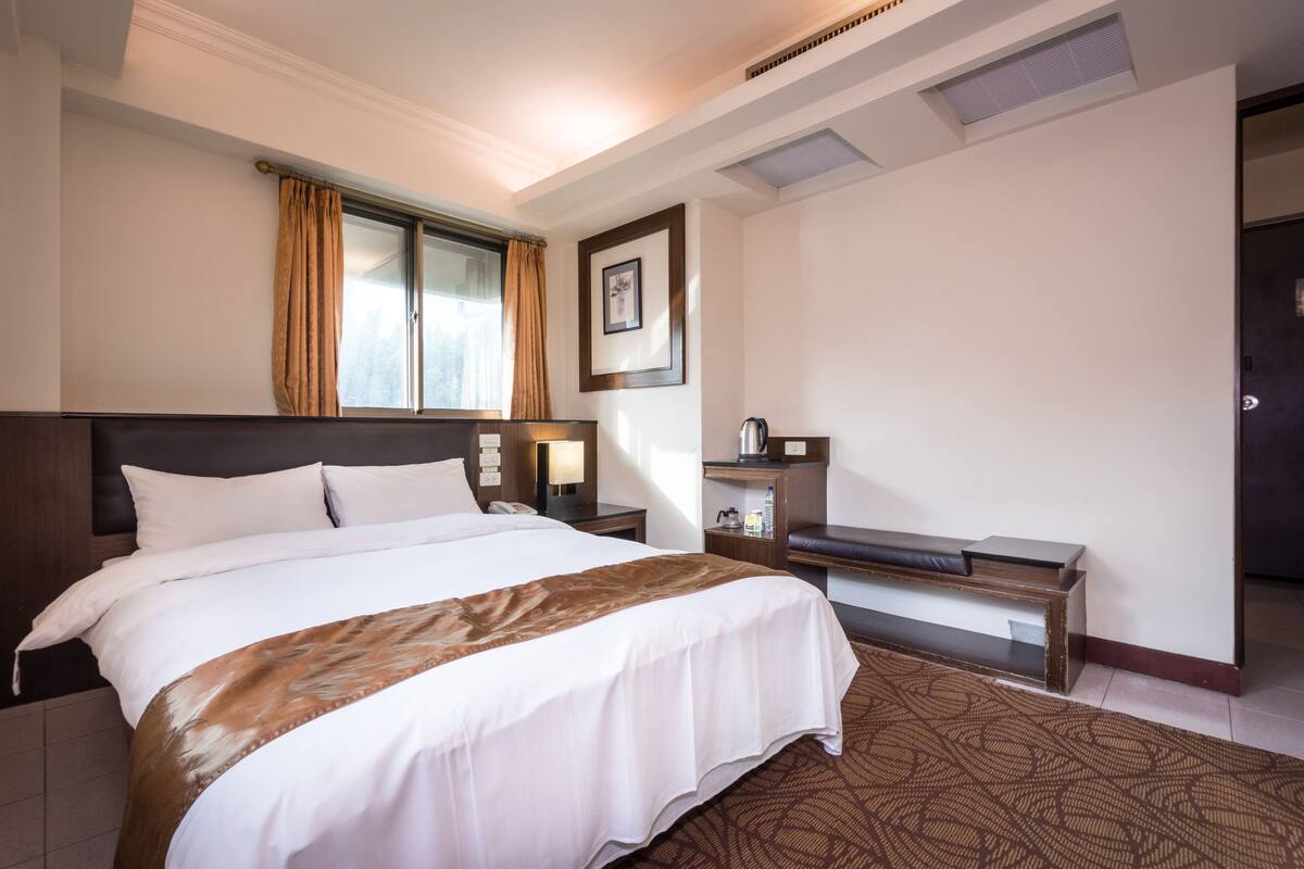 A hotel room with a large bed, a desk, a chair, a nightstand, a lamp, and a framed picture on the wall.