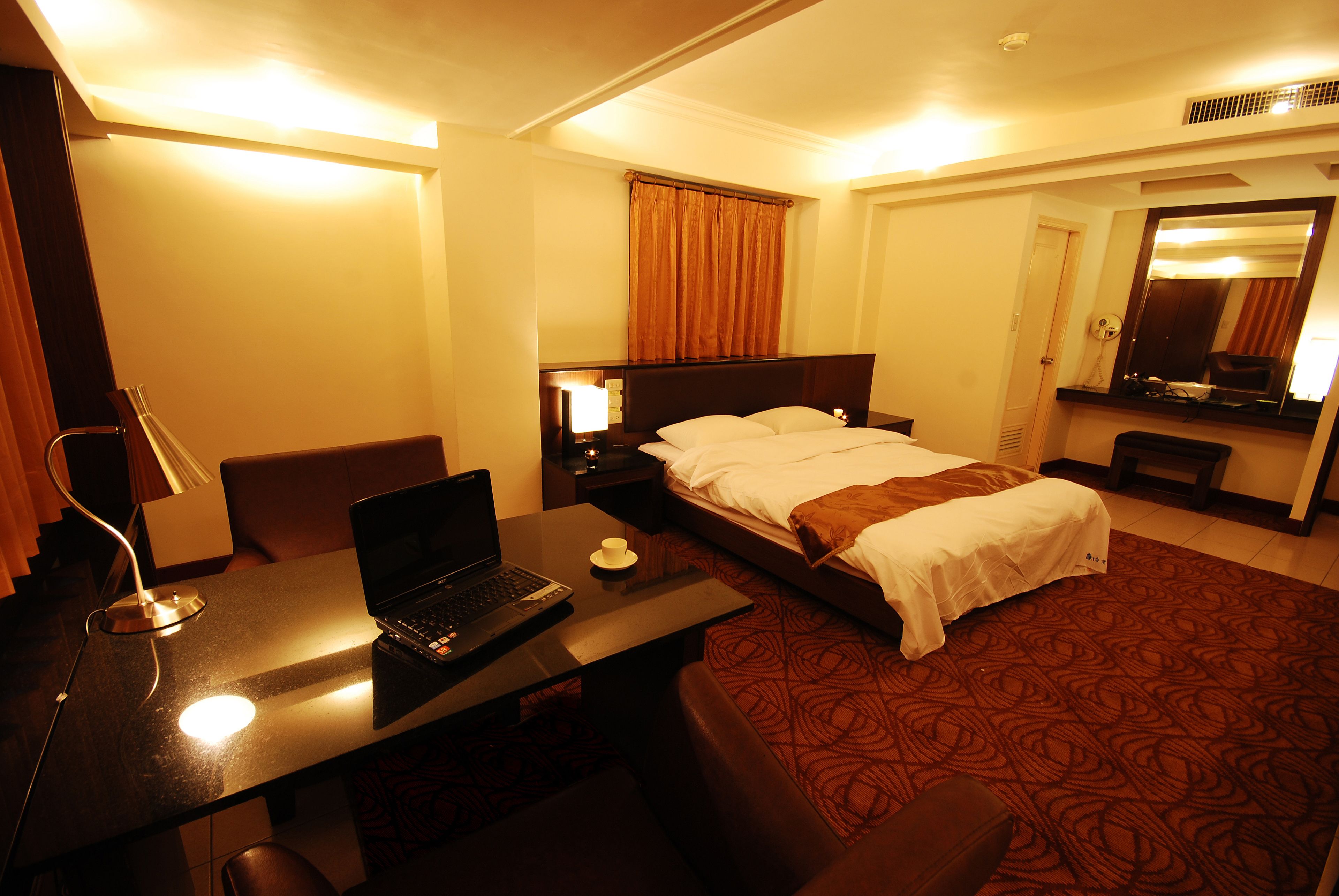 A hotel room with a bed, a desk with a laptop, a chair, a bedside table with a lamp, and a large mirror.