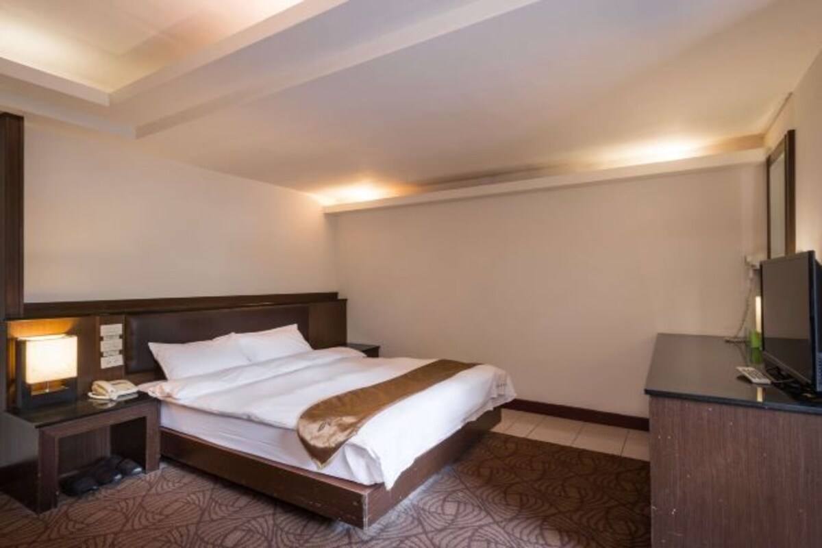 A hotel room with a bed, bedside table, lamp, and a desk with a television.