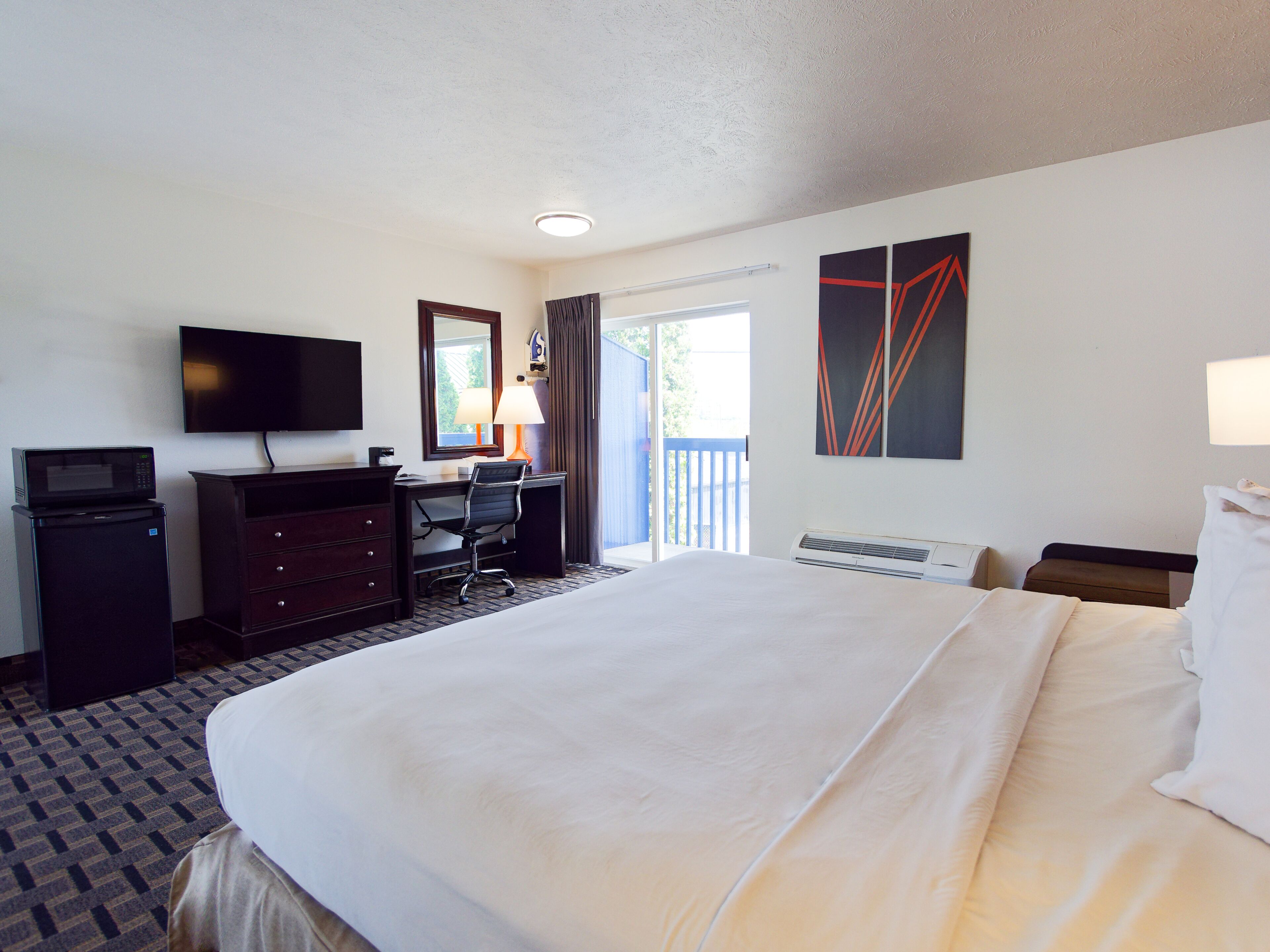 A hotel room with a bed, a desk with a chair, a television, a microwave, a dresser, and a window with curtains.