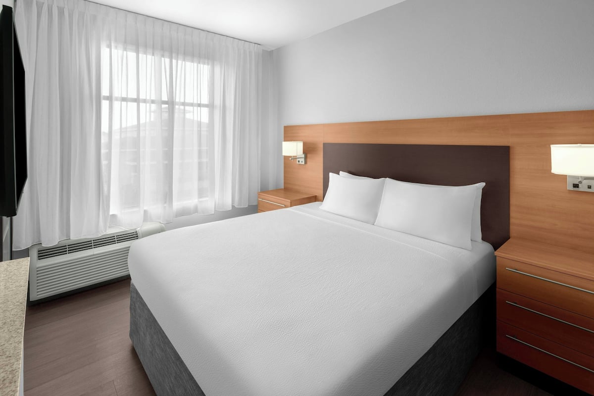 Ett hotellrum med en stor säng, en sänggavel i trä, två sängbord med lampor, ett fönster med tunna gardiner och en väggmonterad luftkonditioneringsenhet.
