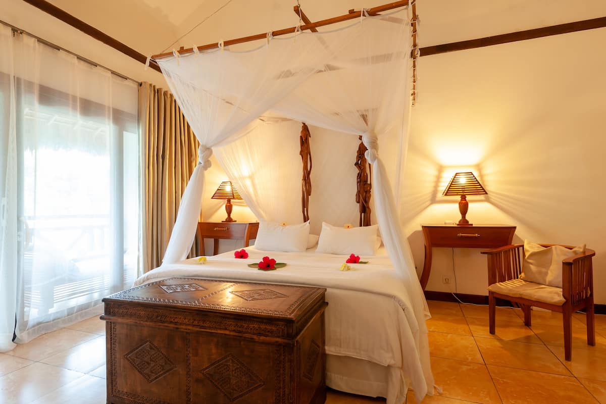 Un lit à baldaquin, deux lampes de chevet, une table de chevet en bois, une chaise en bois et un coffre décoratif en bois.