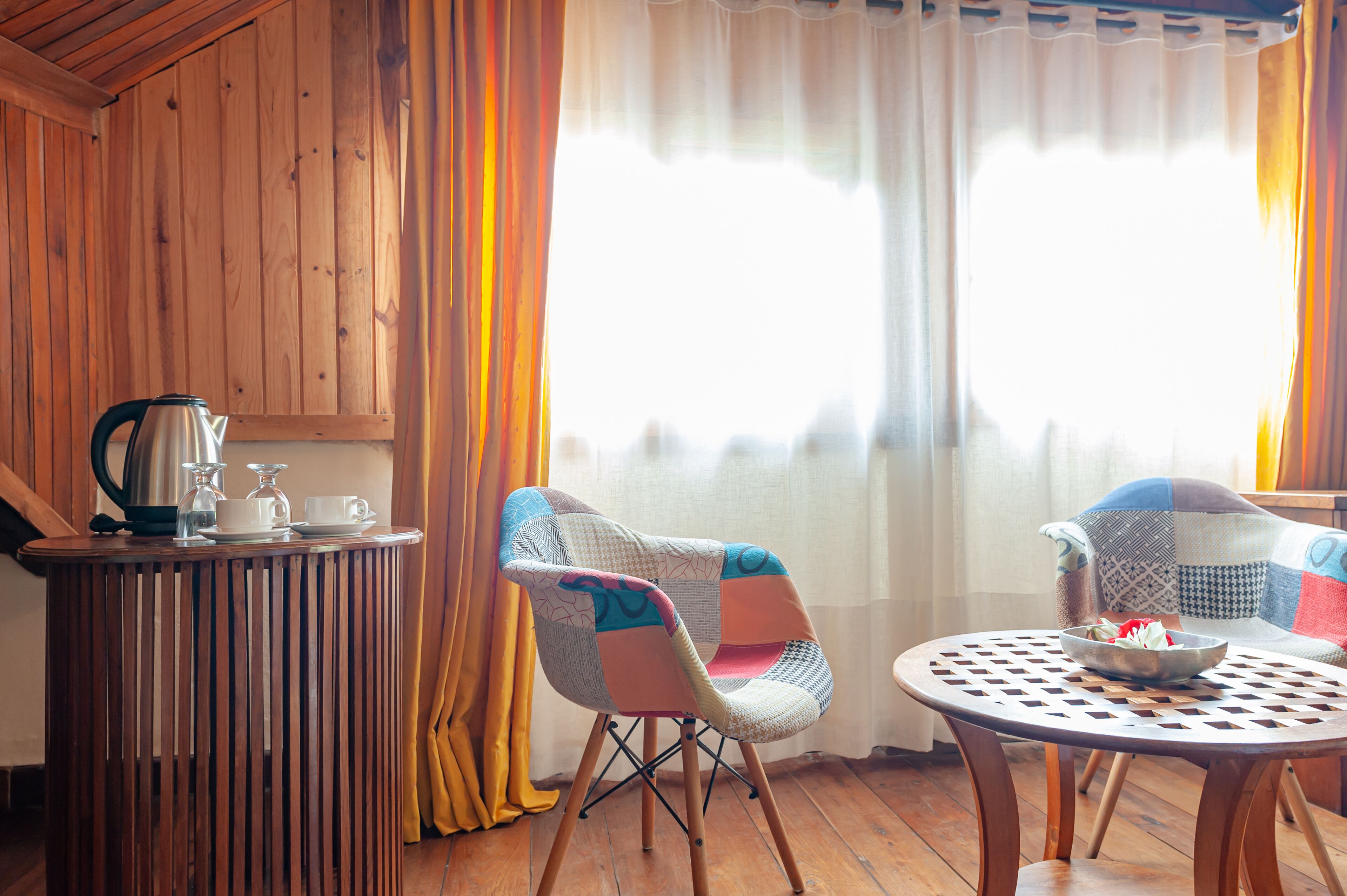 A cozy room with wooden walls, a round table with a bowl, two chairs, and a small table with a kettle and cups.