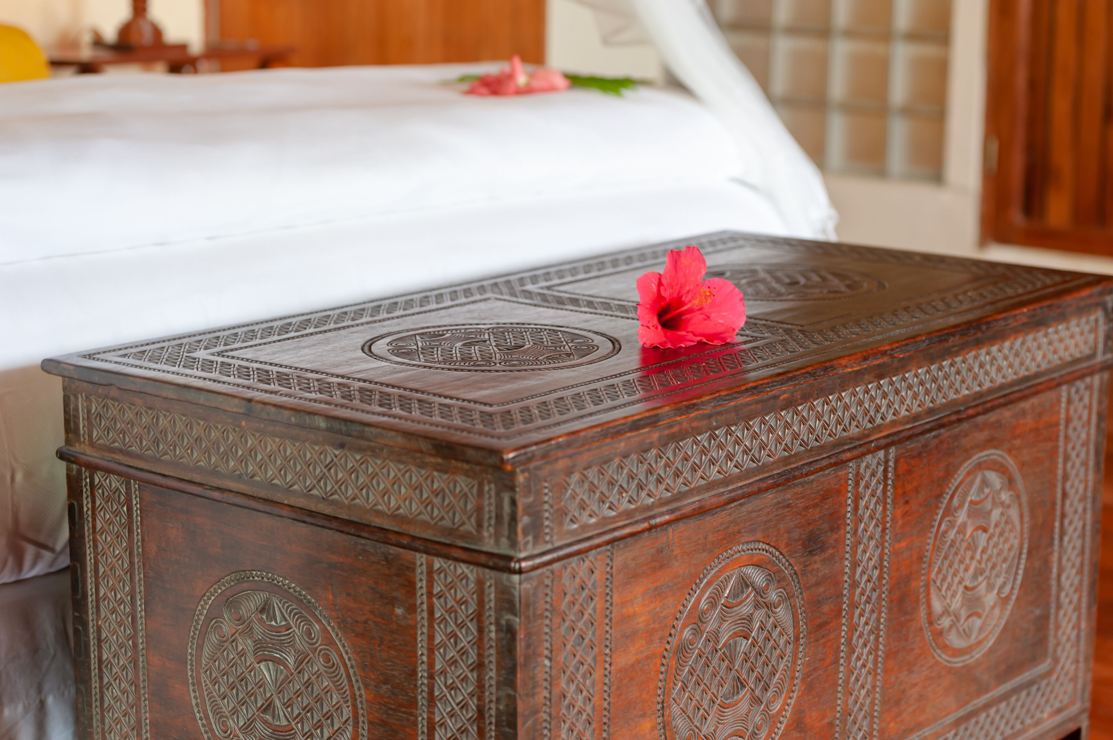 Een houten kist met ingewikkelde gravures en een rode bloem bovenop, geplaatst in een slaapkamer met een bed op de achtergrond.