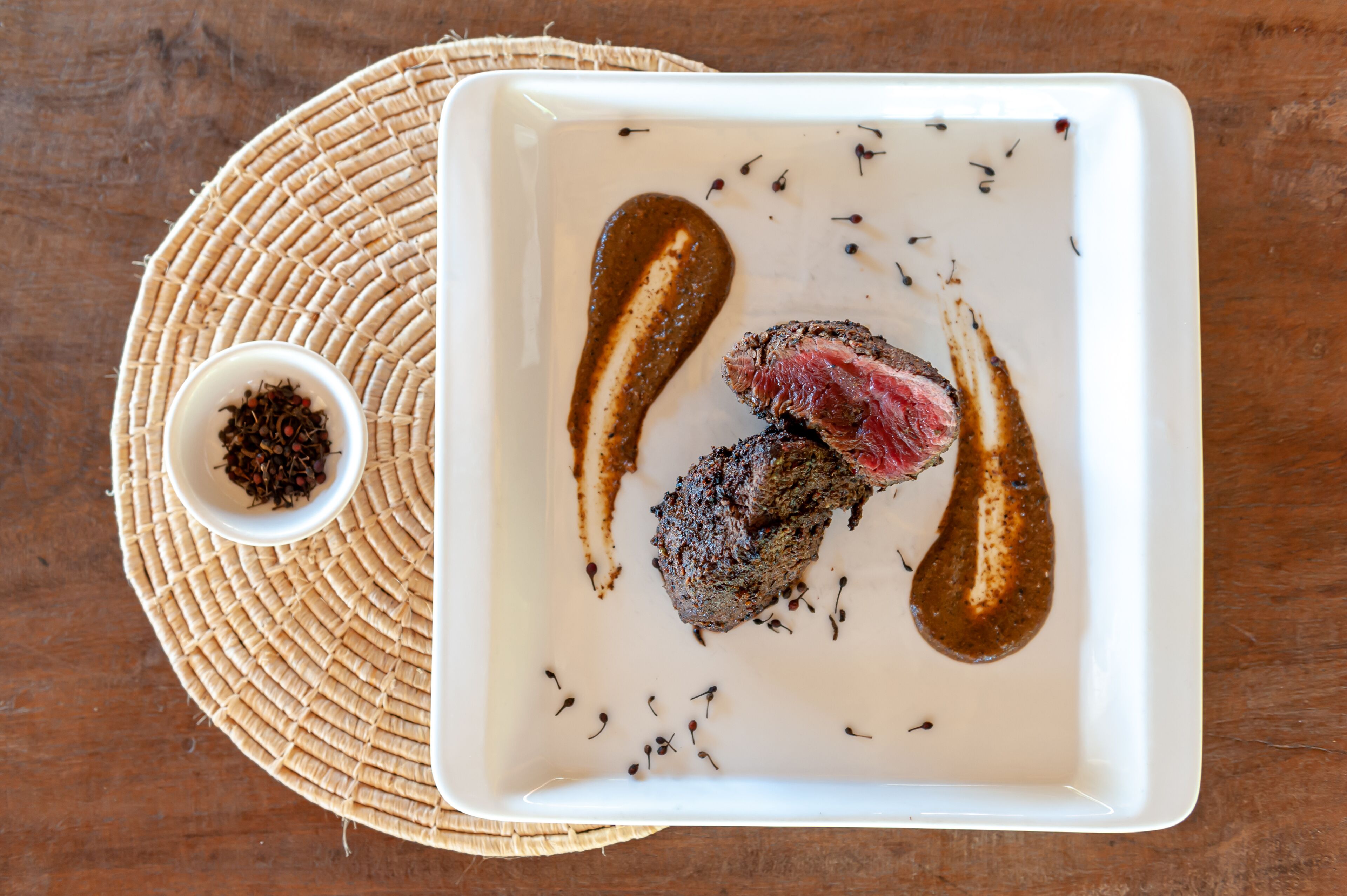 Une assiette carrée blanche sur laquelle on a disposé un steak, une sauce et des herbes, le tout posé sur un set de table en fibres tressées.