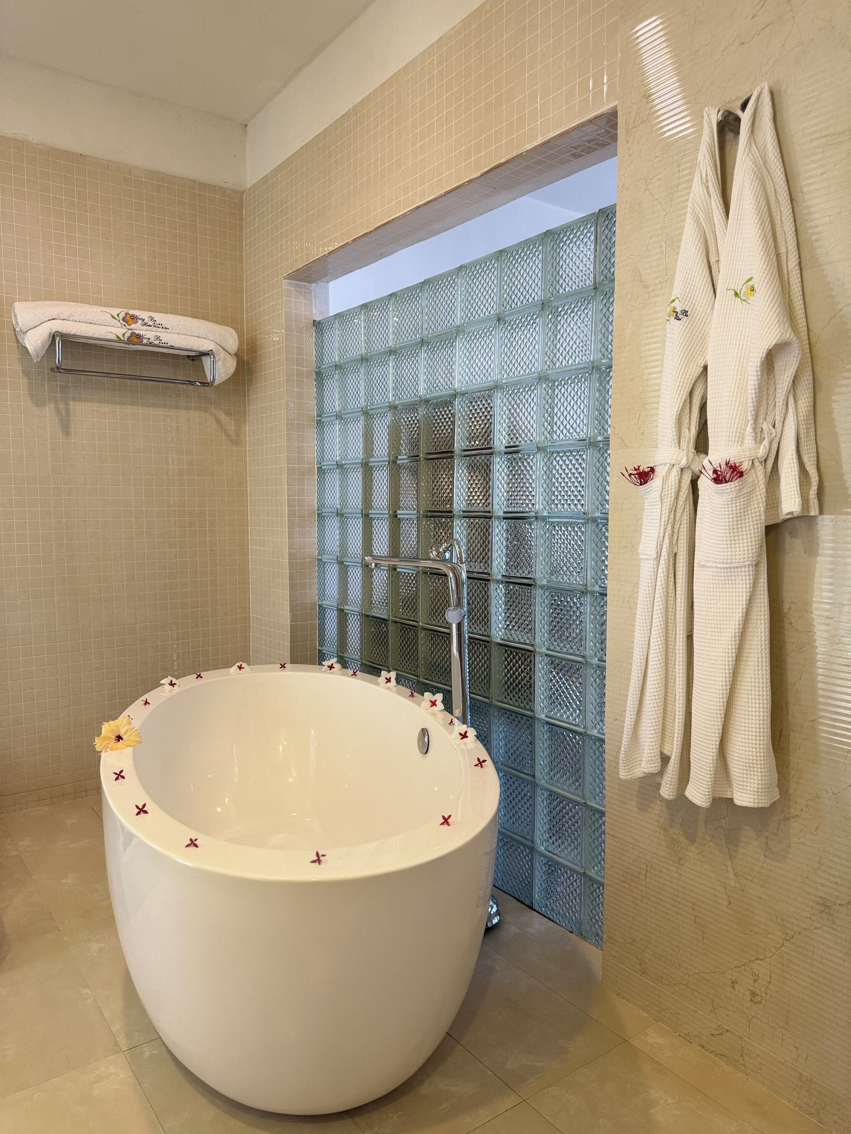 A large freestanding bathtub with a glass enclosure, a towel rack with robes, and a small shelf with a towel.