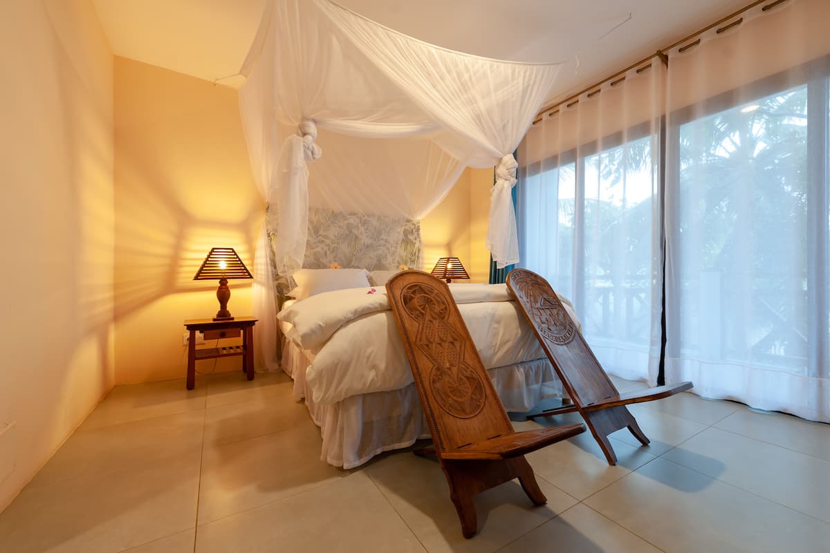 Une chambre à coucher avec un lit à baldaquin, des lampes de chevet, une chaise en bois et une vue sur les palmiers à travers de fines rideaux.