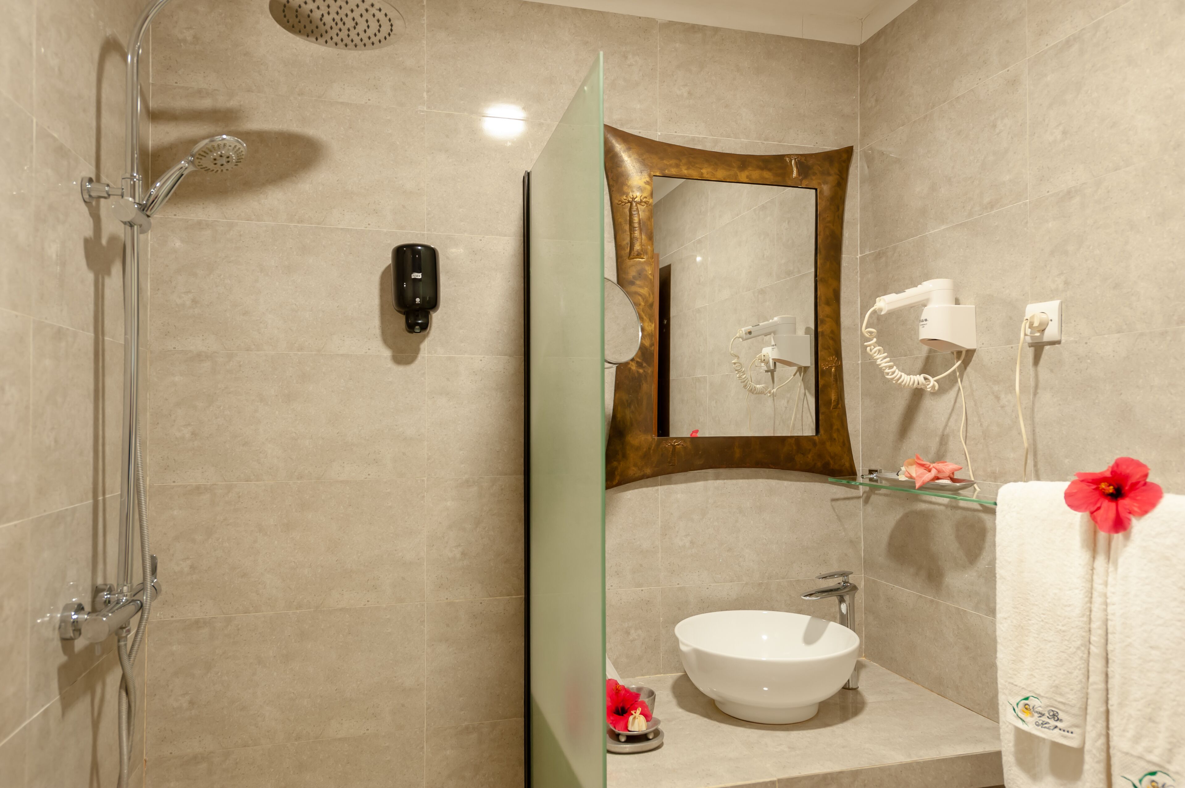 A modern bathroom with a glass shower enclosure, a white sink, a mirror with a gold frame, and a hairdryer on the wall.