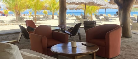 Un salón junto a la playa con techo de paja, asientos acolchados y vista al océano.