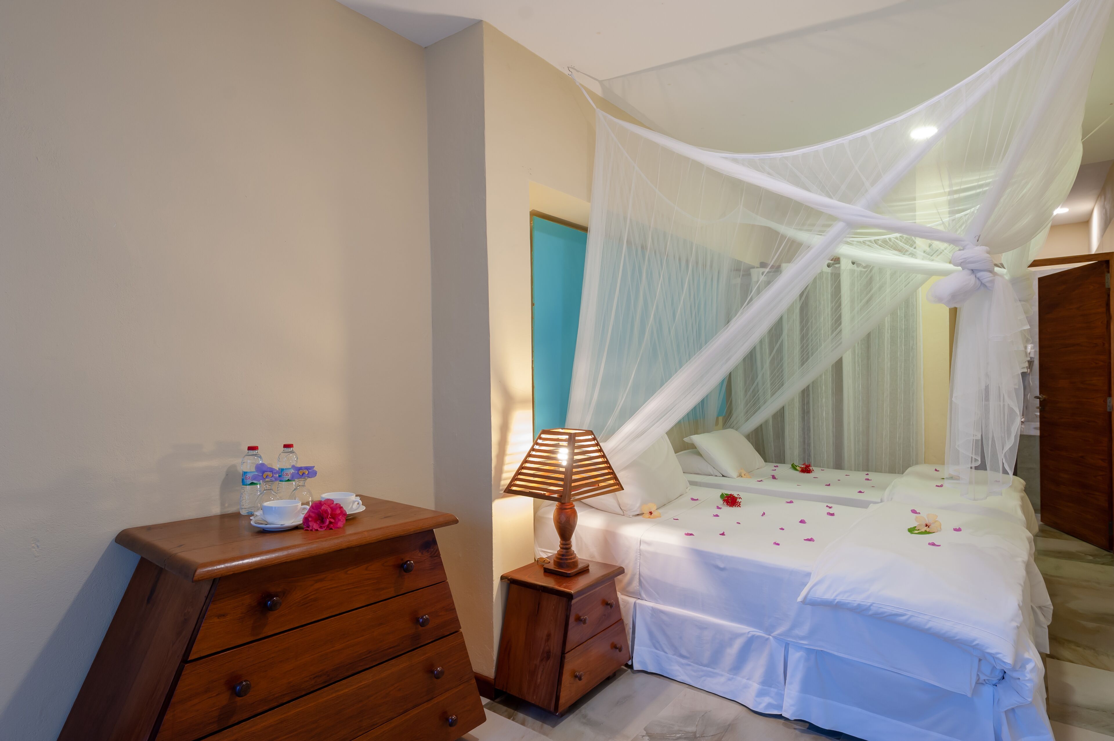 A bedroom with a canopy bed, a wooden nightstand with a lamp, a dresser, and a small table with a water bottle and a flower.