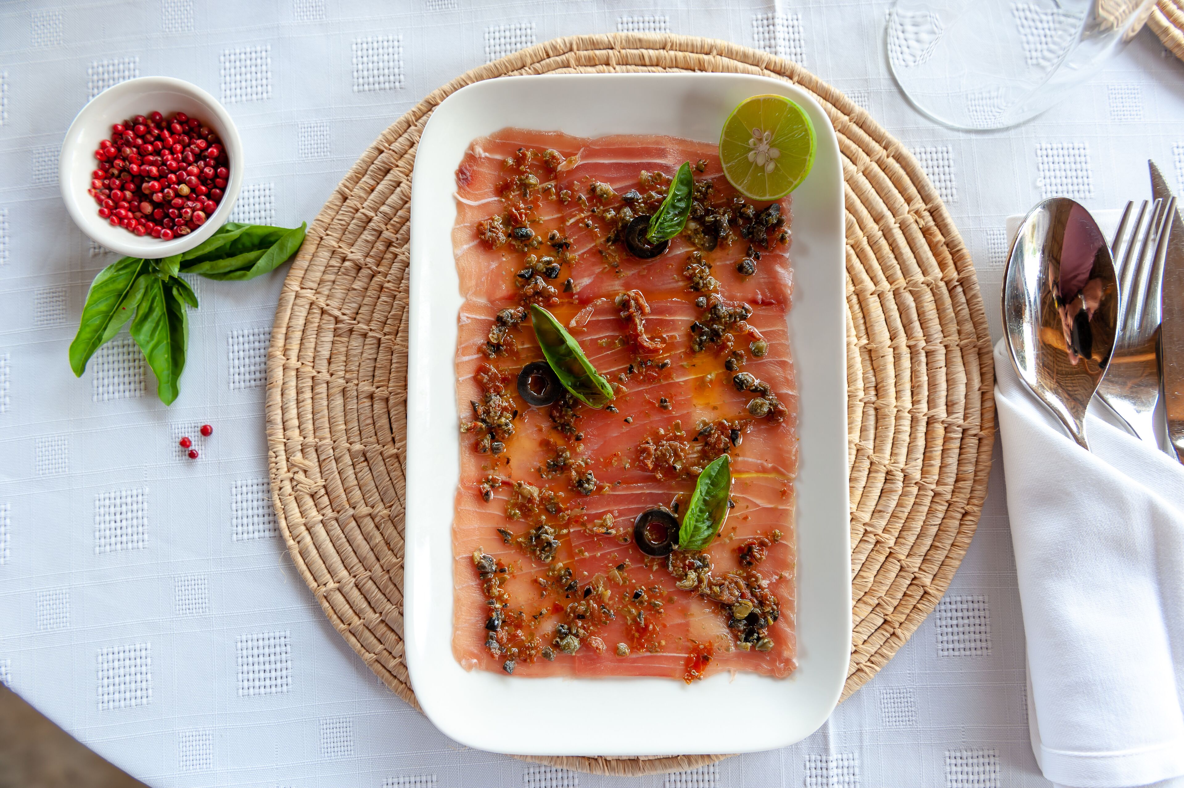 A plate of thinly sliced salmon with capers and a lime wedge, garnished with basil leaves.