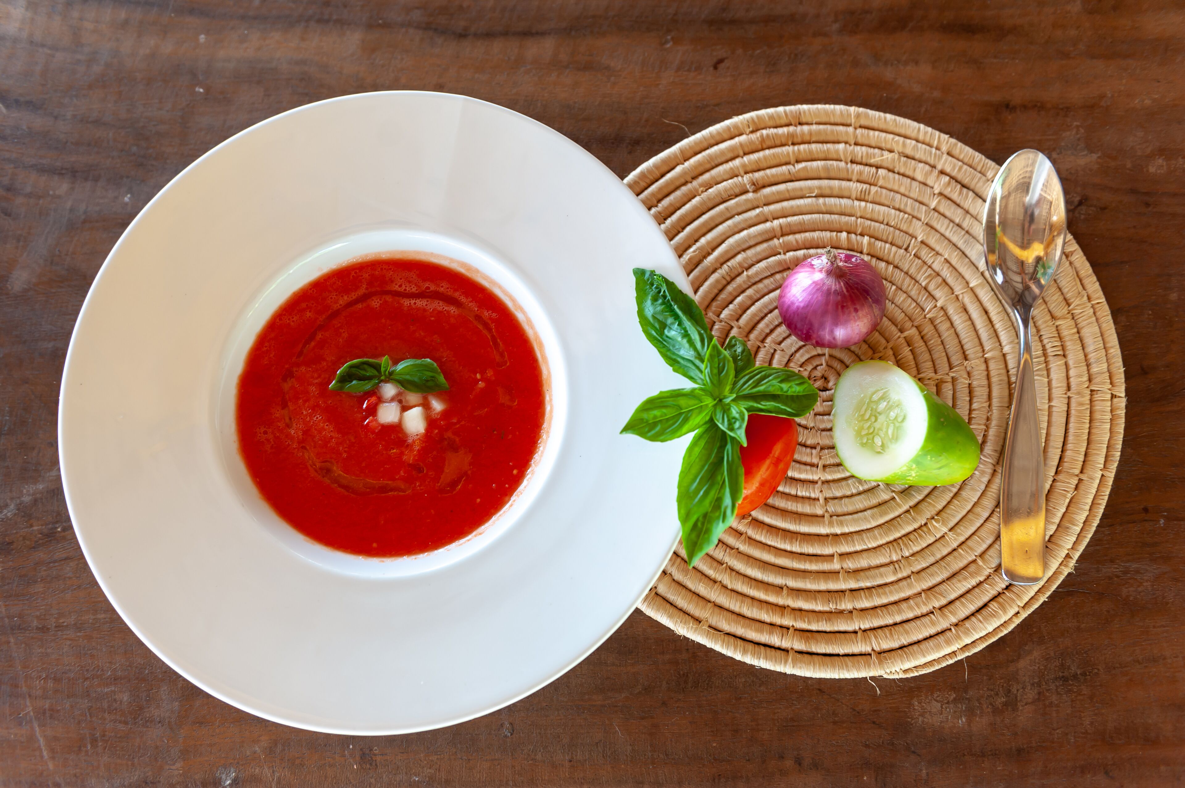 Un tazón de sopa roja sobre un plato blanco, acompañado por una cuchara, un pepino, un tomate y una cebolla roja sobre una estera tejida.