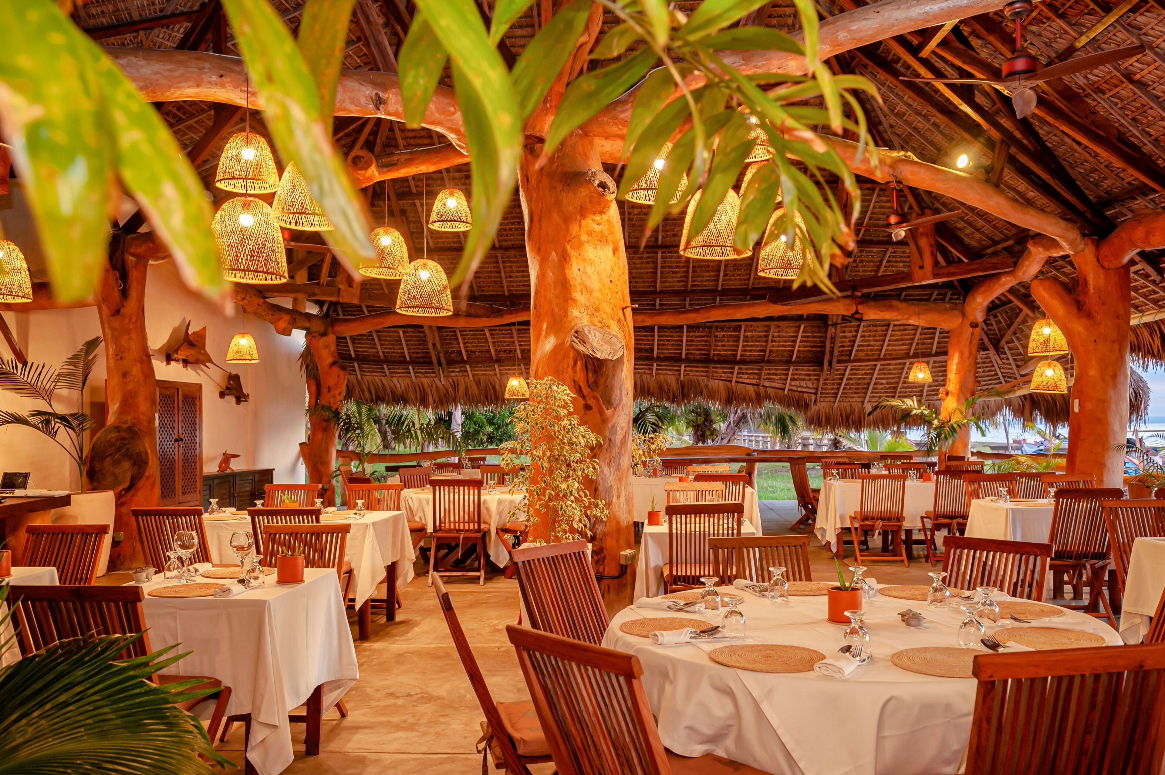 Een restaurantruimte met houten tafels en stoelen, hangende lampen en een rieten dak.