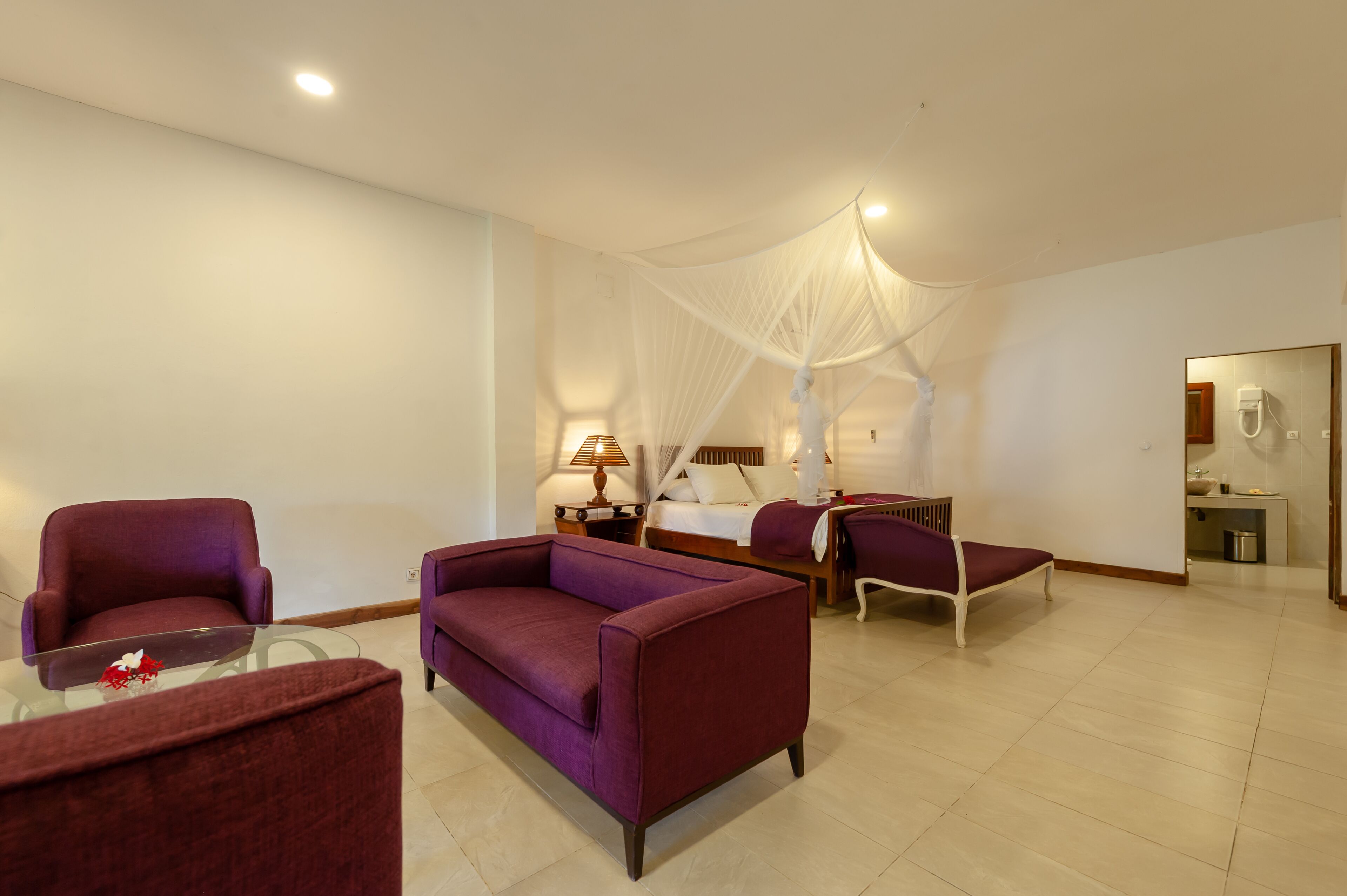A modern hotel room with a large bed, a sitting area with a glass coffee table, and a separate armchair.
