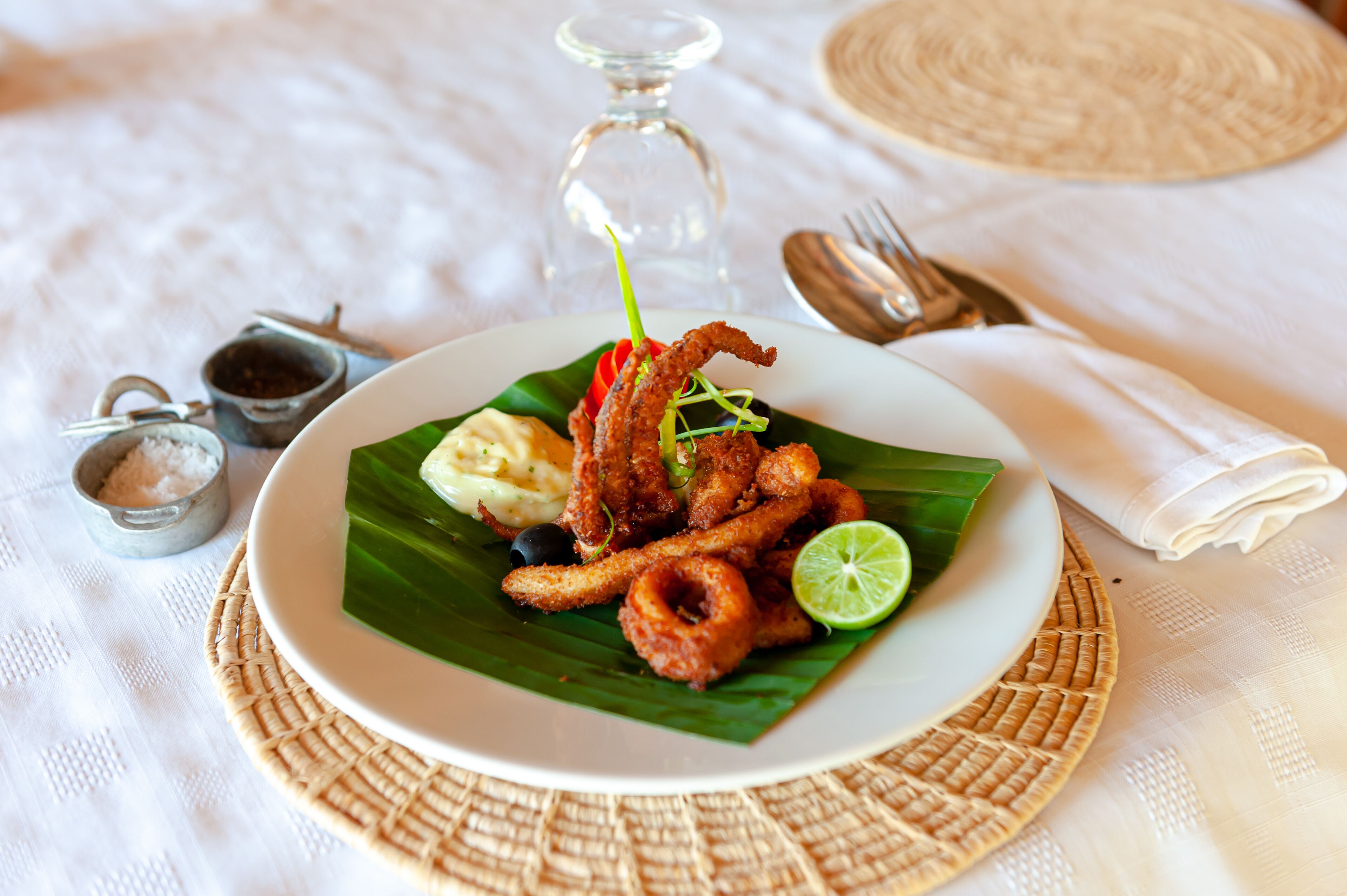A plate of fried seafood on a banana leaf with a lime wedge, accompanied by a small bowl of sauce and a salt shaker.
